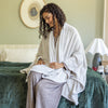 Woman wrapped in a white cape wrap sitting on a bed in a bedroom setting.