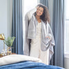Woman wrapped in a gray cape wrap standing in a bedroom with curtains and a bed.