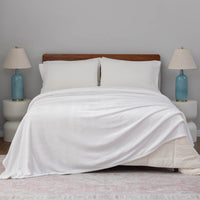 A white velvet blanket draped over a bed with white pillows and blue lamps.