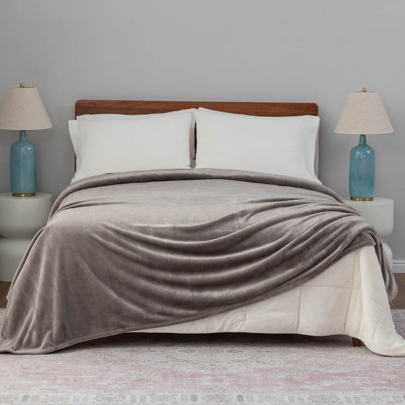 A gray velvet blanket draped over a bed with white pillows and blue lamps.