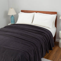 Polartec Softec Microfleece blanket in black draped on a bed, with white pillows and a blue lamp on the side.