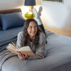 Woman laying on a bed with a polar blue Polartec Softec Microfleece blanket, as she smiles and reads a book.