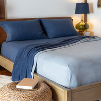 Bedroom scene with a Polartec Softec Microfleece blanket in light blue, draped over a bed next to a wicker end table with books and a navy glass lamp.