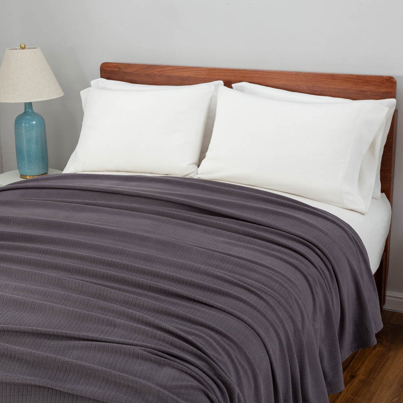 Polartec blanket in dark gray draped over a bed with white pillows and a blue lamp.