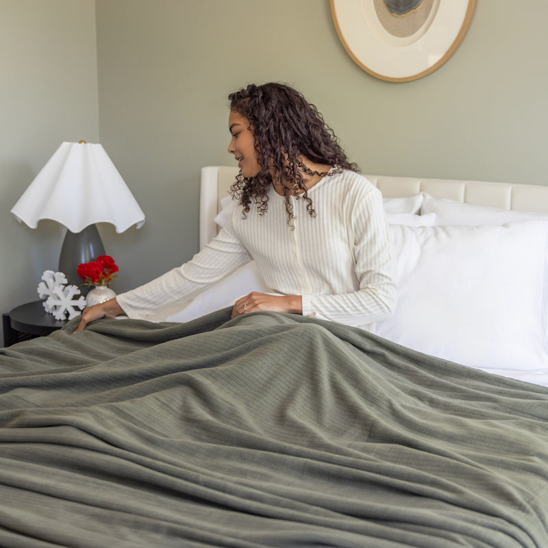 Woman in a white sweater arranging a green blanket on a bed in a bedroom.