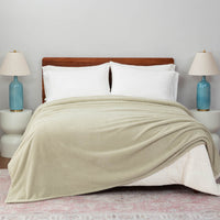 A Retro Comfort Plush Blanket in sage green draped over a bed with white pillows and light blue bedside lamps.