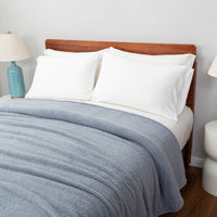 A minimalist bedroom with a blue Retro Comfort Plush Blanket on a bed, accompanied by a wooden bed frame and blue lamp.