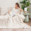 A woman sitting on a beige sofa with a neutral botanical faux fur heated throw draped over her legs as she reads a book.