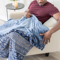 A close up of a blue fairisle printed heated throw laying on a man's legs as he sits on a chair.