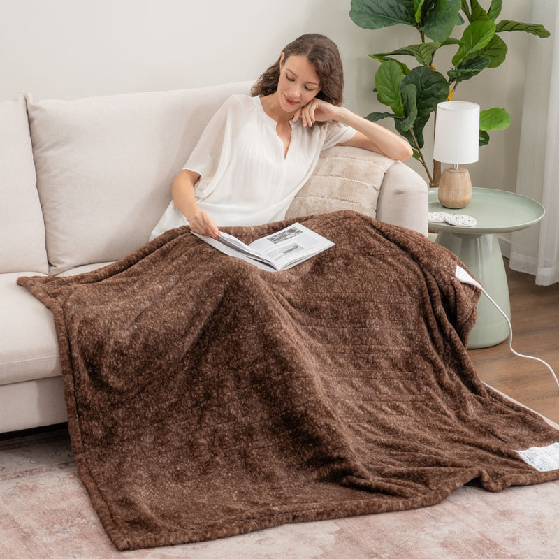 A woman sitting on a beige sofa with a brown faux fur heated throw draped over her legs as she reads a book.