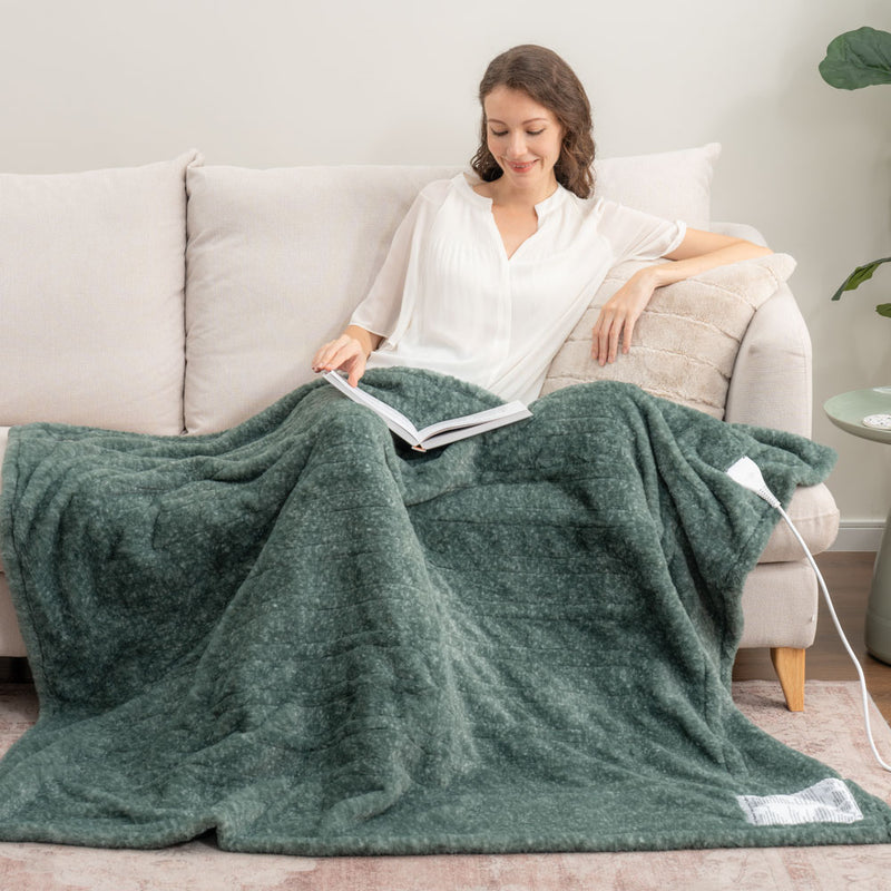 A woman sitting on a beige sofa with a green faux fur heated throw draped over her legs as she reads a book.