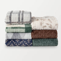 A stack of seven folded heated throws in a variety of prints and colors on a white background.