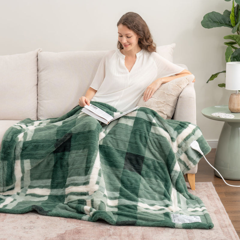 A woman sitting on a beige sofa with a green plaid faux fur heated throw draped over her legs as she reads a book.