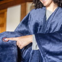 Woman wearing a navy heated throw, standing in a room while pressing the power button.