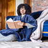 Woman leaning on a blue couch wearing a navy heated wearable throw, as she flips through a book.