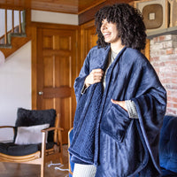 Woman wearing a navy heated wearable throw, standing in front of a wooden door by a chair.