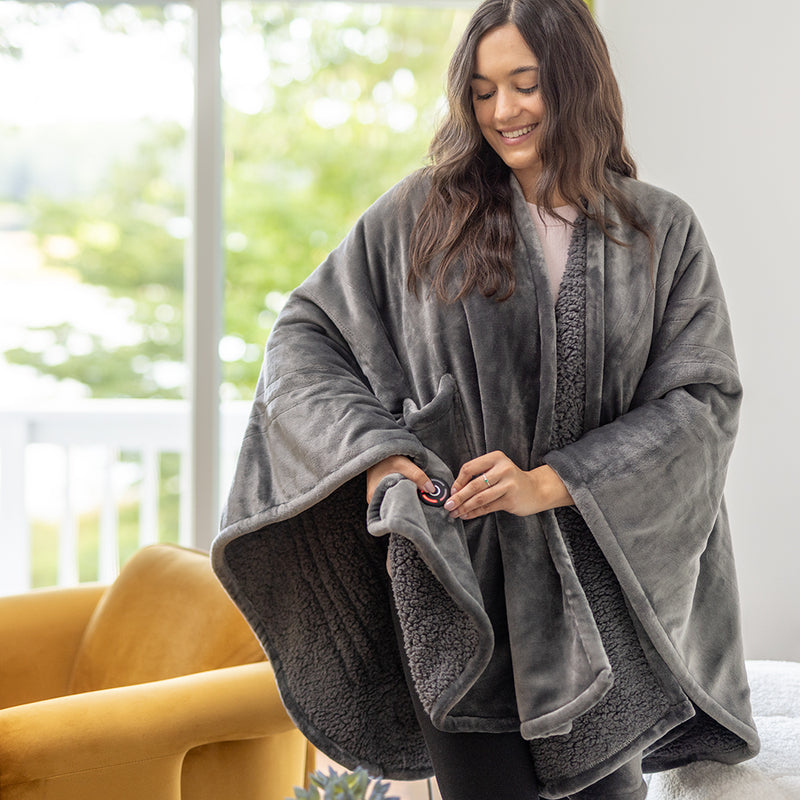 Woman in a gray heated wearable throw, seated on a couch with a cozy, relaxed posture.