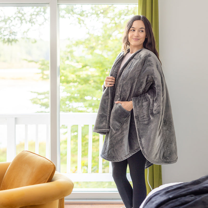 Woman in a gray heated wearable throw, standing in front of a window.