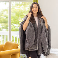 Woman modeling a grey heated wearable throw as she stands in front of a window.