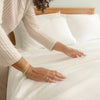 Person adjusting white bedding on a bed with a neutral background