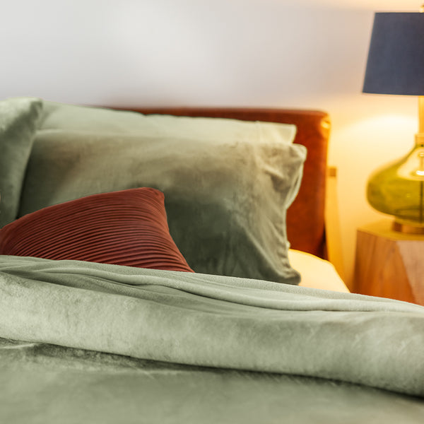 Close-up of a bed styled with a Dark Sage VelvetLoft® duvet cover and pillow shams, accented by a rust-colored decorative pillow. A warm bedside lamp with a green glass base softly illuminates the scene.