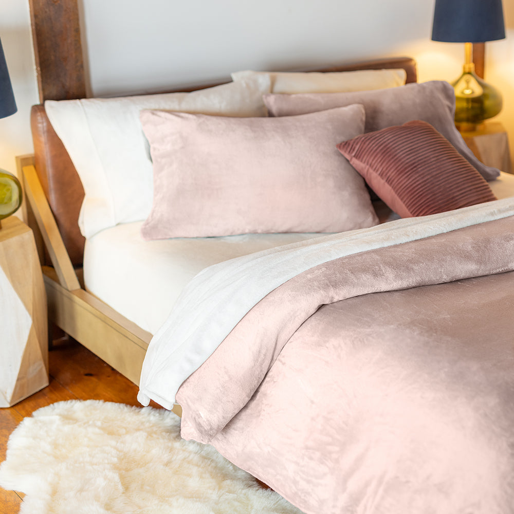 Bed styled with a Rose Smoke VelvetLoft® duvet cover and pillow shams, accented by a rust-colored decorative pillow. A green glass lamp and a soft cream rug add warmth and elegance to the bedroom setting.