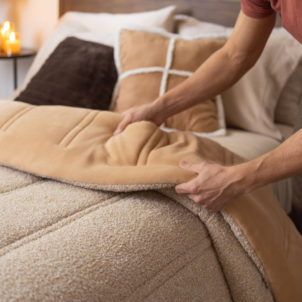 A person making a bed, adjusting a soft tan comforter with a cozy sherpa lining. Behind the bed, decorative pillows in neutral shades of beige and brown add warmth to the scene. A lit candle glows softly in the background, creating a calming and inviting atmosphere.