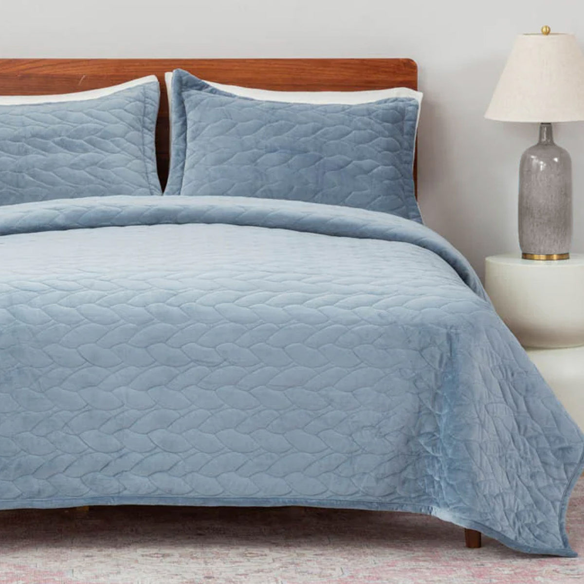 Blue quilted bedspread on a bed with wooden headboard and lamp in the background.
