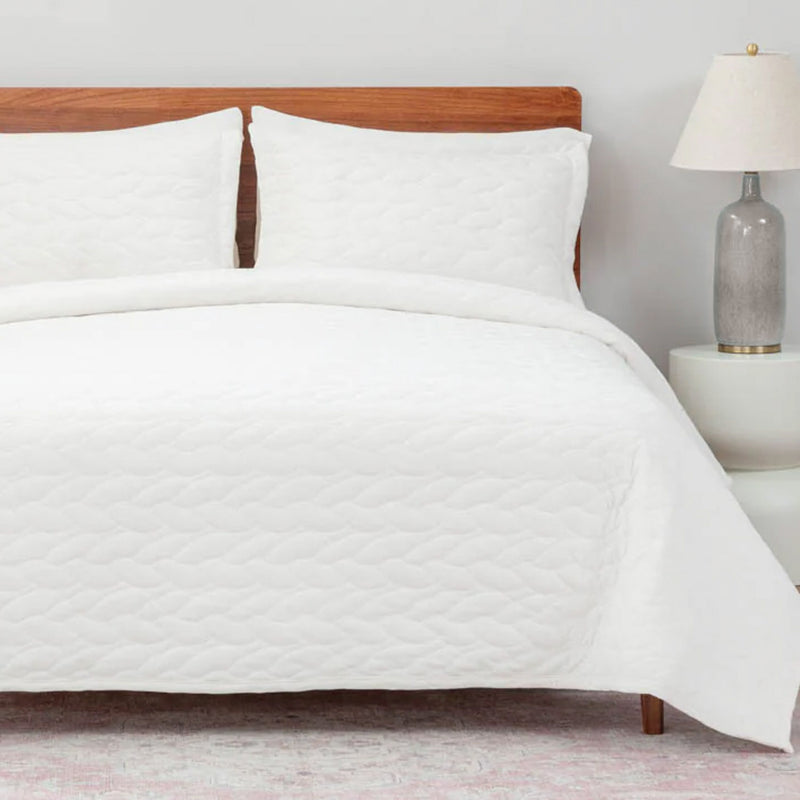 White quilted comforter on a bed with wooden headboard and lamp in the background