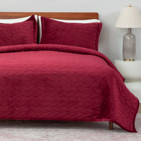 Red quilted bedspread and pillows on a bed with a lamp in the corner.