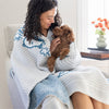 A woman sitting on a chair holding a dog with our Peanuts MellowSoft Throw wrapped around her shoulders.