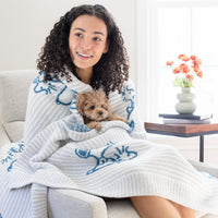 A woman sitting on a chair holding a dog with our Peanuts MellowSoft Throw wrapped around her shoulders.