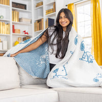 A woman sitting on a sofa with our Peanuts MellowSoft Throw wrapped around her shoulders.