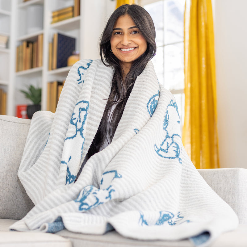 A woman sitting on a sofa with our Peanuts MellowSoft Throw wrapped around her shoulders.