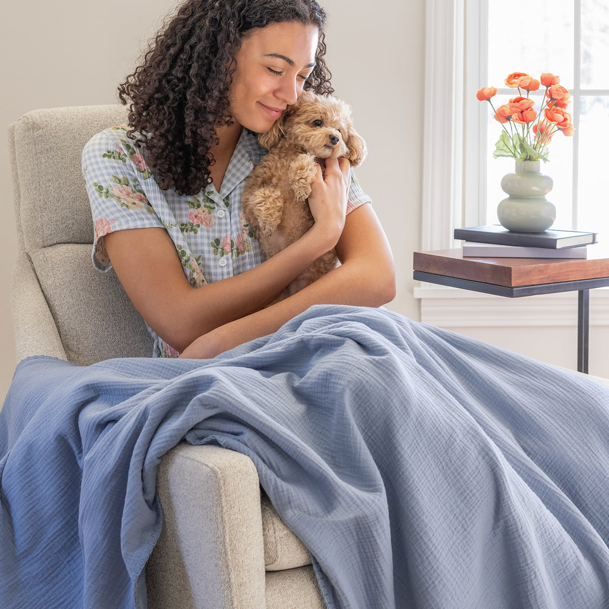 A woman holding a dog while sitting on  beige chair with a blue gauze throw.