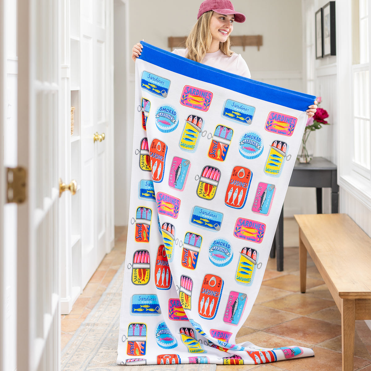 A woman holding up a sardines beach towel in a bright room.