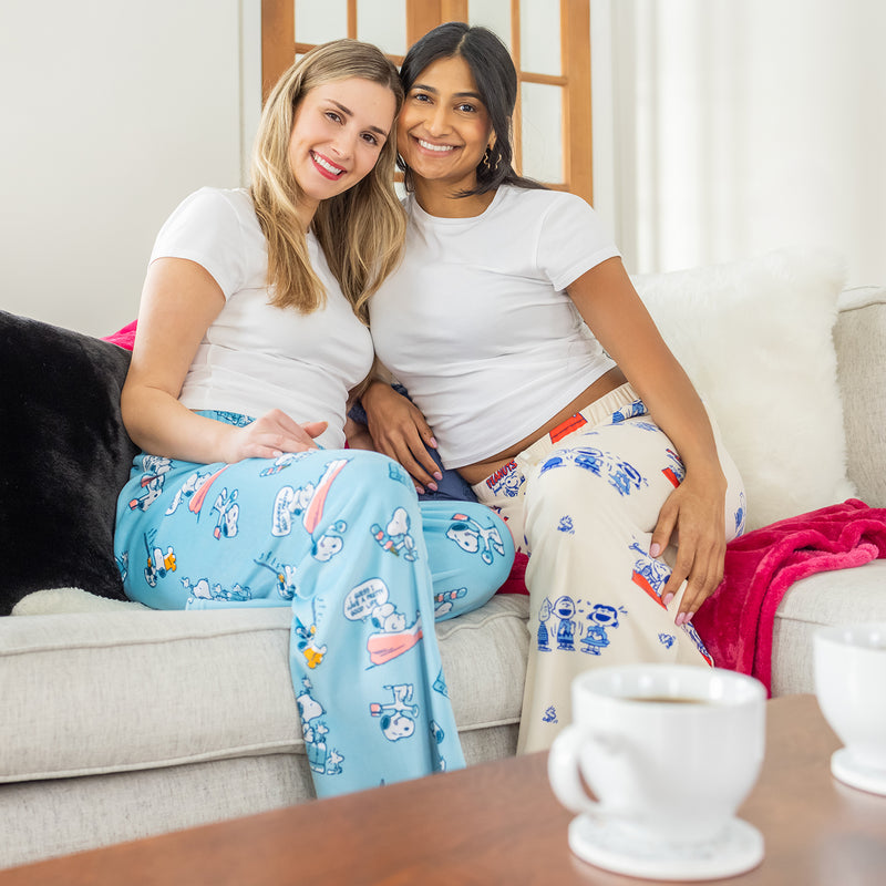 Two woman wearing Peanuts printed lounge pants while sitting next to eachother on a sofa.
