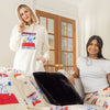 A woman wearing a cream Peanuts lounge hoodie standing next to a woman sitting on a sofa wearing Peanuts lounge pants.