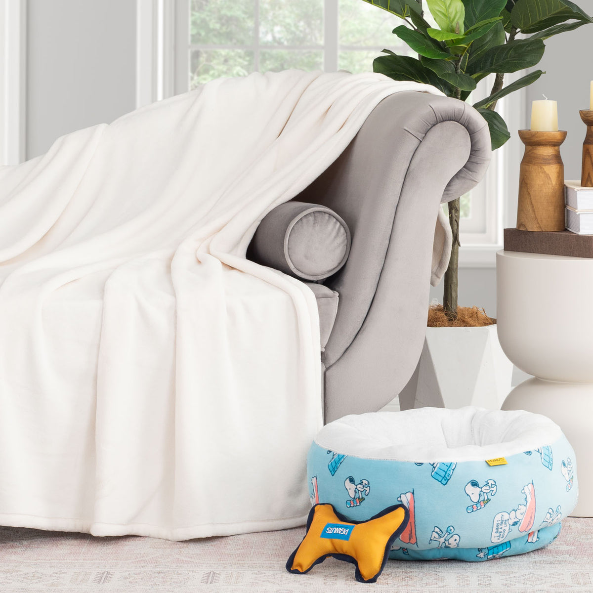 Peanuts pet bed and dog toy set sitting next to a sofa and cream throw in a staged room.