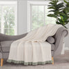 White blanket with green trim draped over a gray sofa in a room with large windows and a plant.