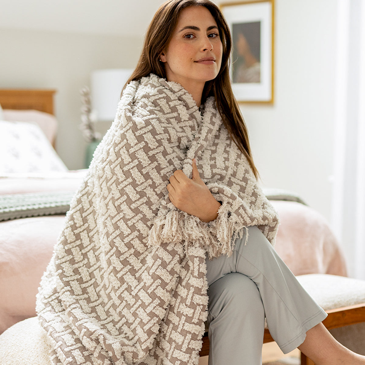 Woman wrapped in a patterned blanket sitting on a bench in bedroom with bed in the background. 