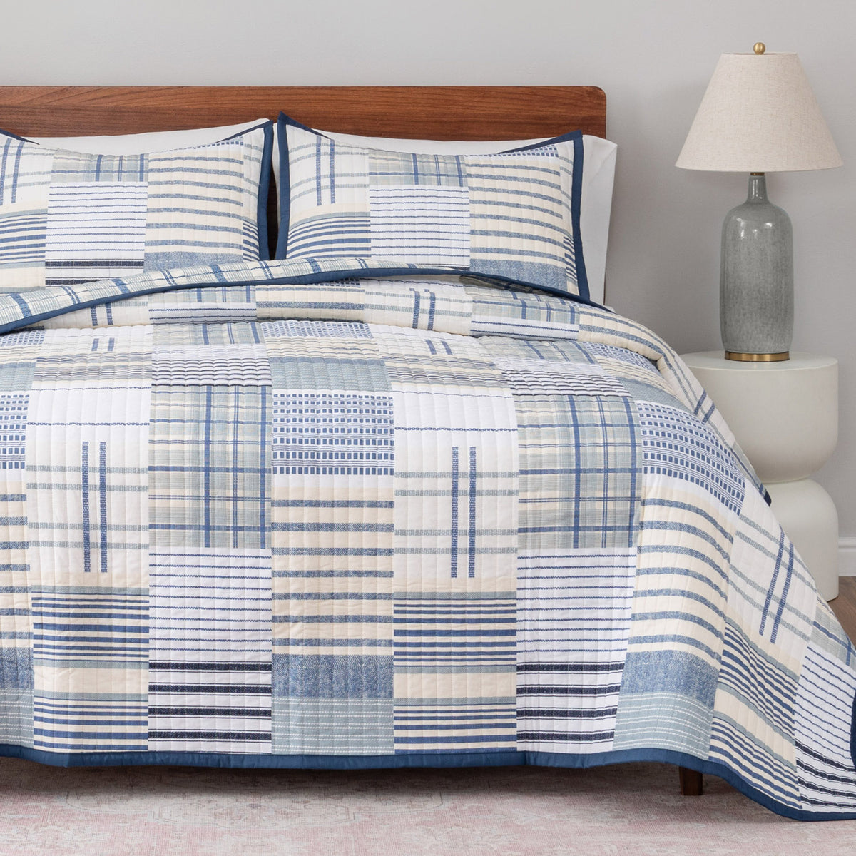 Bed with blue and white checkered comforter and pillows, nightstand with lamp in the background.