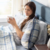 Woman sitting up in bed with a stripe quilted blanket and a mug, looking relaxed.