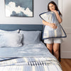 Woman holding a striped quilted pillowcase in a bedroom with a bed and artwork on the wall.