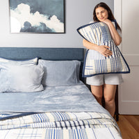 Woman holding a striped quilted pillowcase in a bedroom with a bed and artwork on the wall.