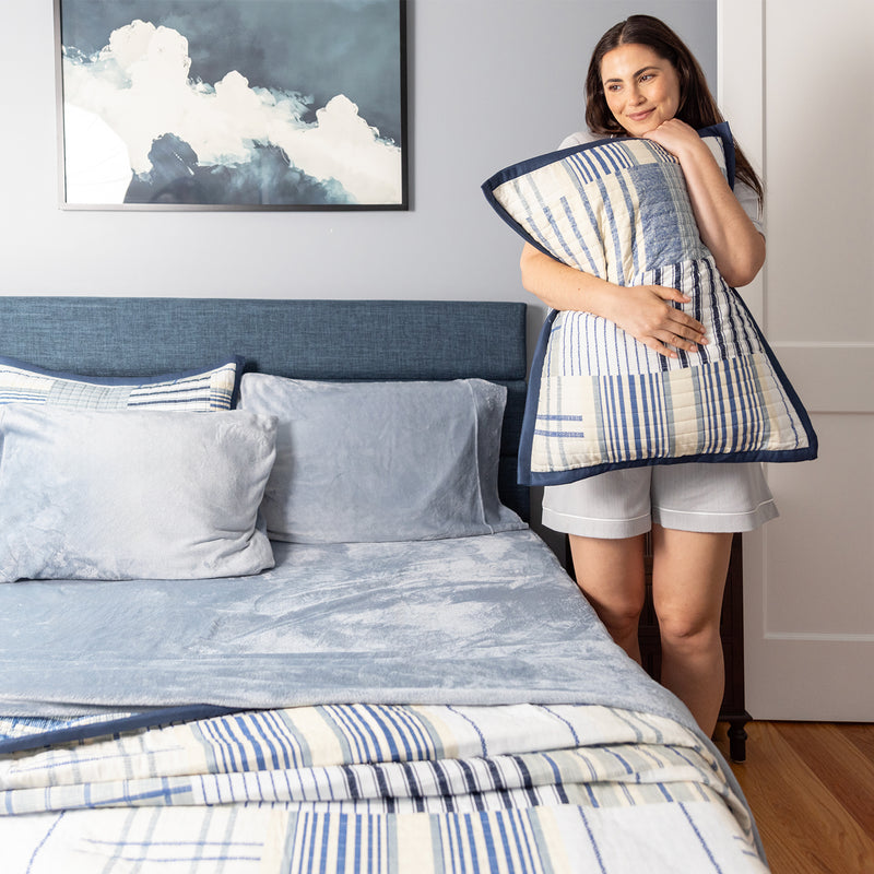 Woman holding a striped quilted pillowcase in a bedroom with a bed and artwork on the wall.