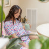 Woman in a floral robe holding a green mug with a straw, sitting on a couch.
