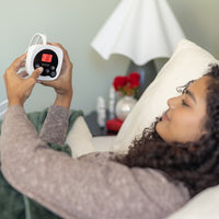 Woman lying on a bed holding a digital thermometer with a blurred room background