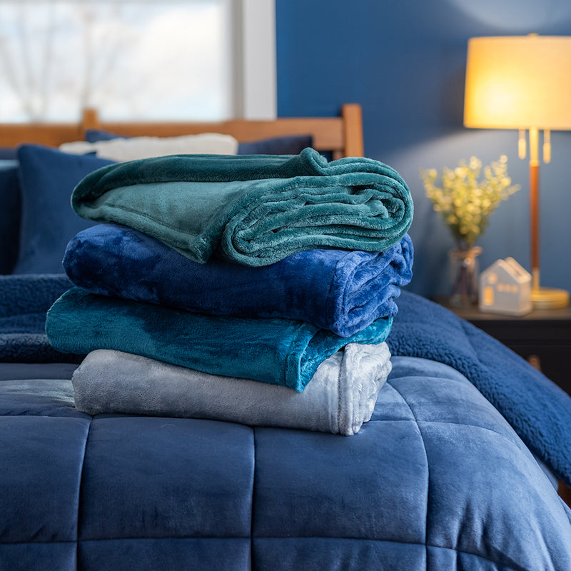 A stack of 4 blue VelvetLoft Throws stacked on a blue comforter in a cozy bedroom.