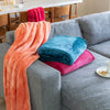 Our blue and pink VelvetLoft Throws stacked on a grey couch with our orange VelvetLoft Throw draped over the arm of the sofa.